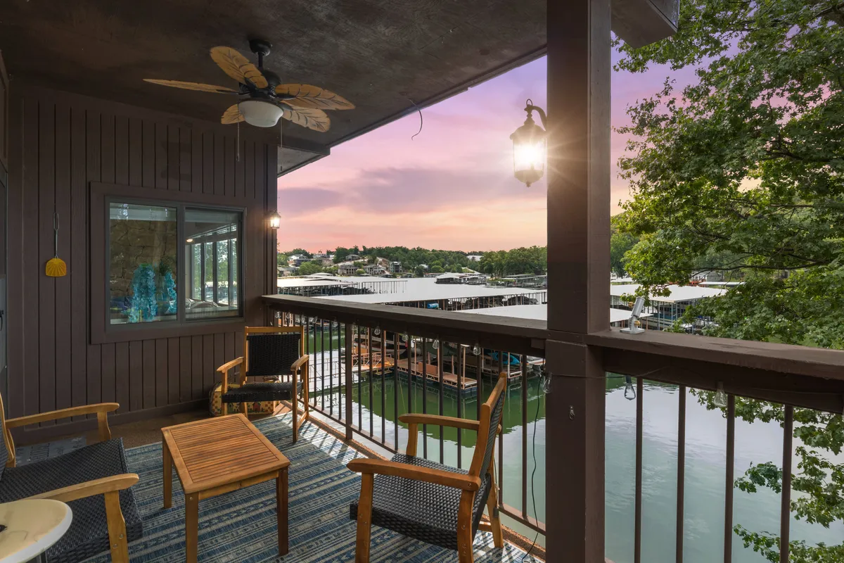 The hero image shows a serene exterior patio with comfortable wooden chairs and a small table, all under a cooling ceiling fan. The patio overlooks a breathtaking lake view, with the sun setting in the distance, casting warm colors across the water. The space is designed for relaxing, enjoying a drink, or sharing conversation while immersed in tranquil natural beauty.