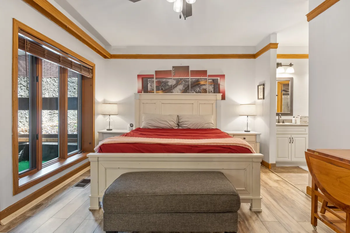 A serene bedroom featuring a bed with red bedding, artwork above the bed, and large windows that bathe the room in natural light. The space feels cozy, restful, and thoughtfully decorated for comfort.