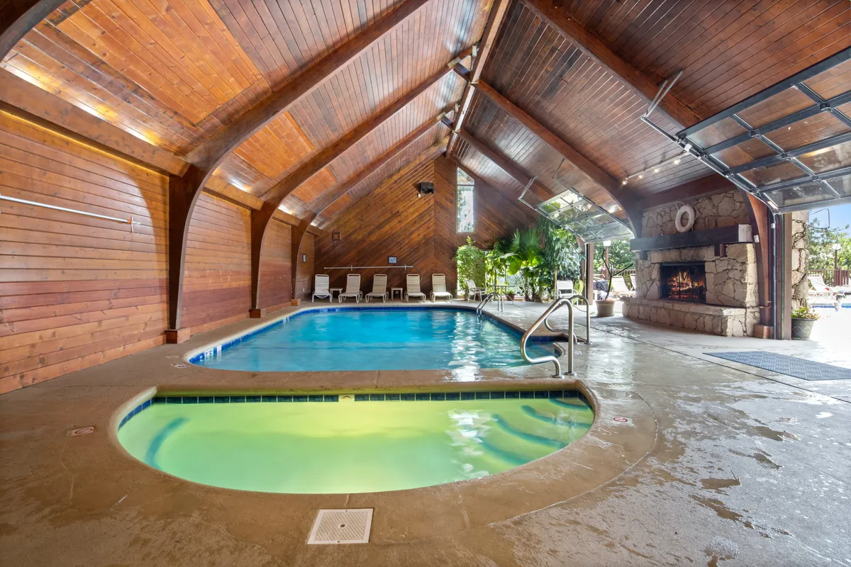 An interior scene featuring an indoor pool and adjacent spa area with a wooden ceiling and a cozy fireplace. The lighting is soft and inviting, emphasizing luxury and relaxation. The space is ideal for swimming, socializing, or simply unwinding in a resort-like atmosphere.