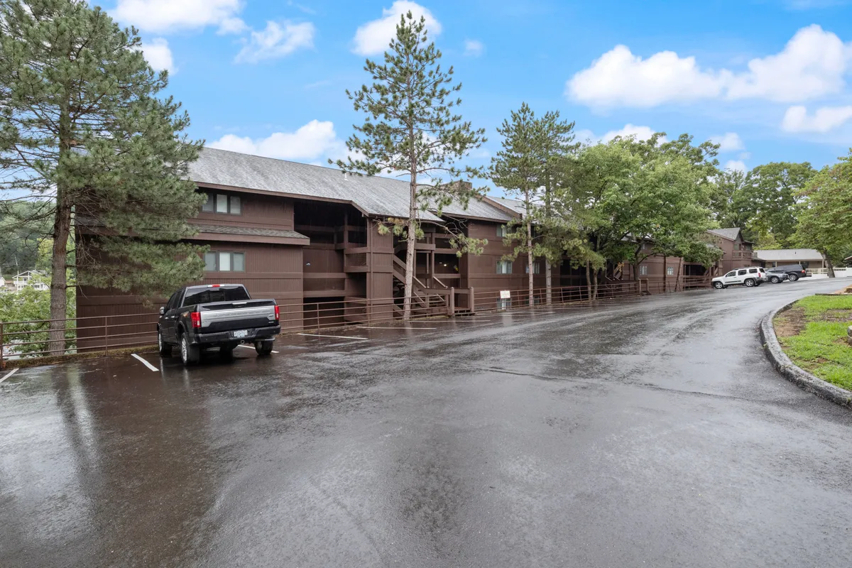 This exterior image features a peaceful parking lot bordered by mature trees and the building's facade, suggesting a tranquil arrival experience and a strong connection to nature. The trees and gentle outdoor setting evoke the idea of stepping into a serene retreat, which aligns with the section’s emphasis on relaxation and natural surroundings.