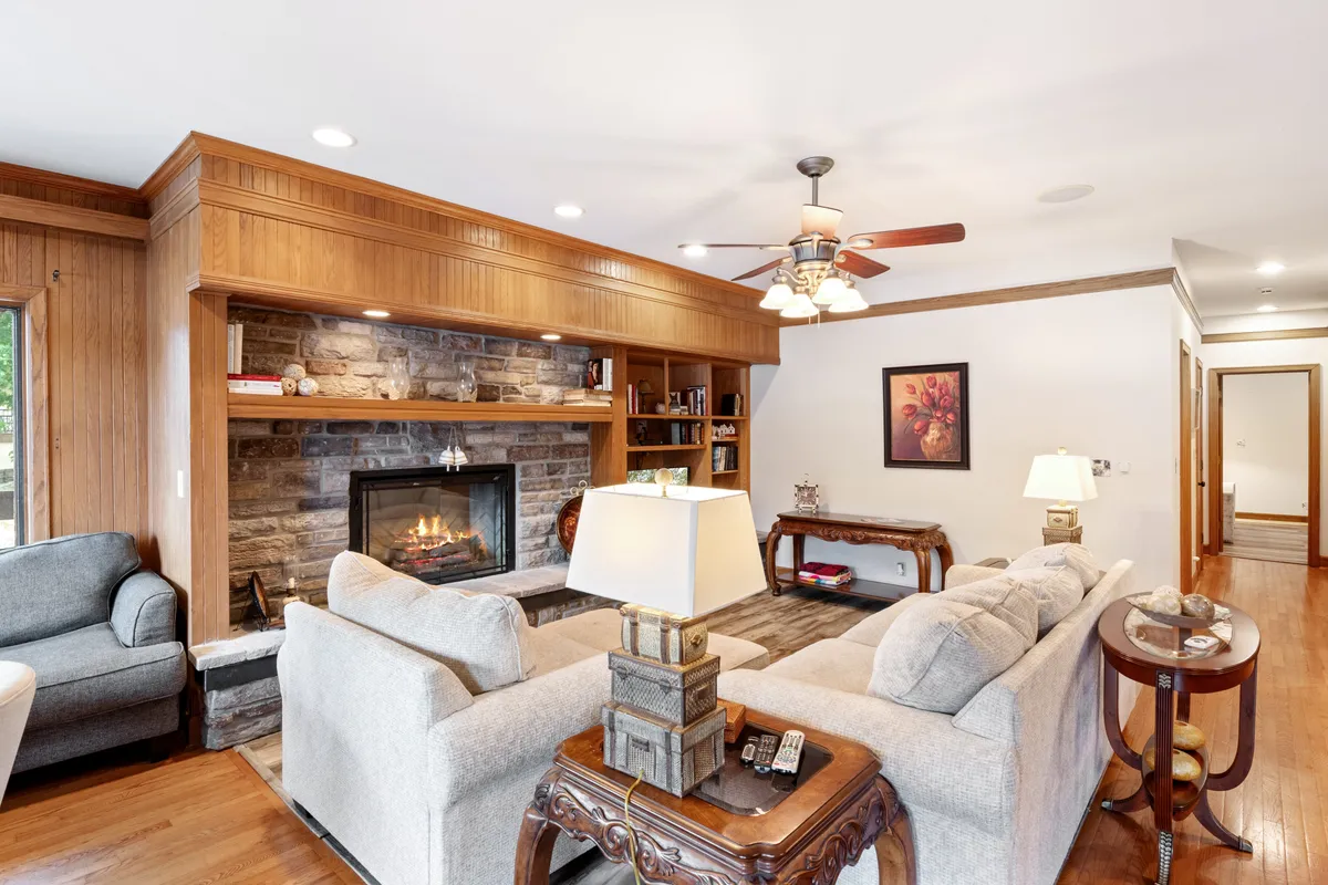 A warm, inviting living room featuring a decorative fireplace framed by elegant woodwork and stonework. A comfortable sofa, bookshelves, and a softly lit lamp complete the cozy ambiance, suggesting a perfect setting for relaxation, family gatherings, or curling up with a good book.