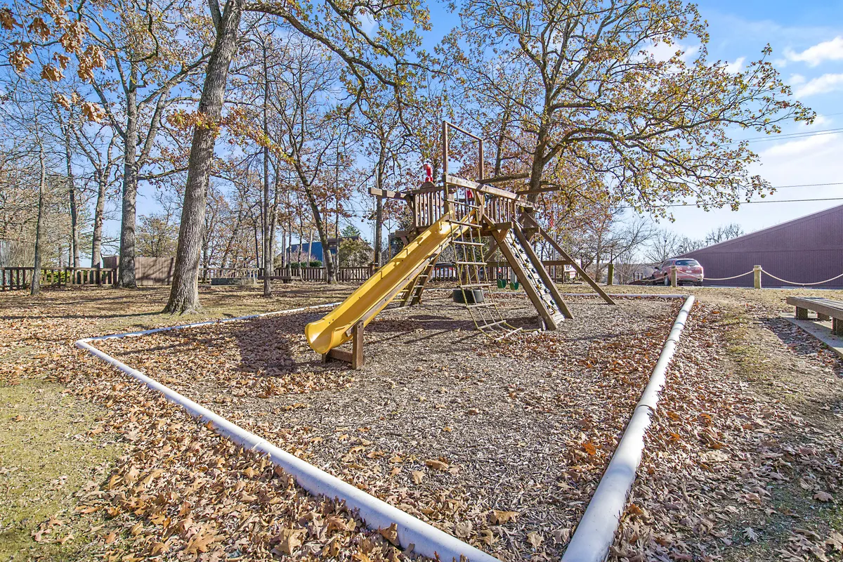 An exterior playground scene surrounded by trees, showing children playing on slides and swings. The space is vibrant and designed for family enjoyment, community gatherings, and outdoor fun.