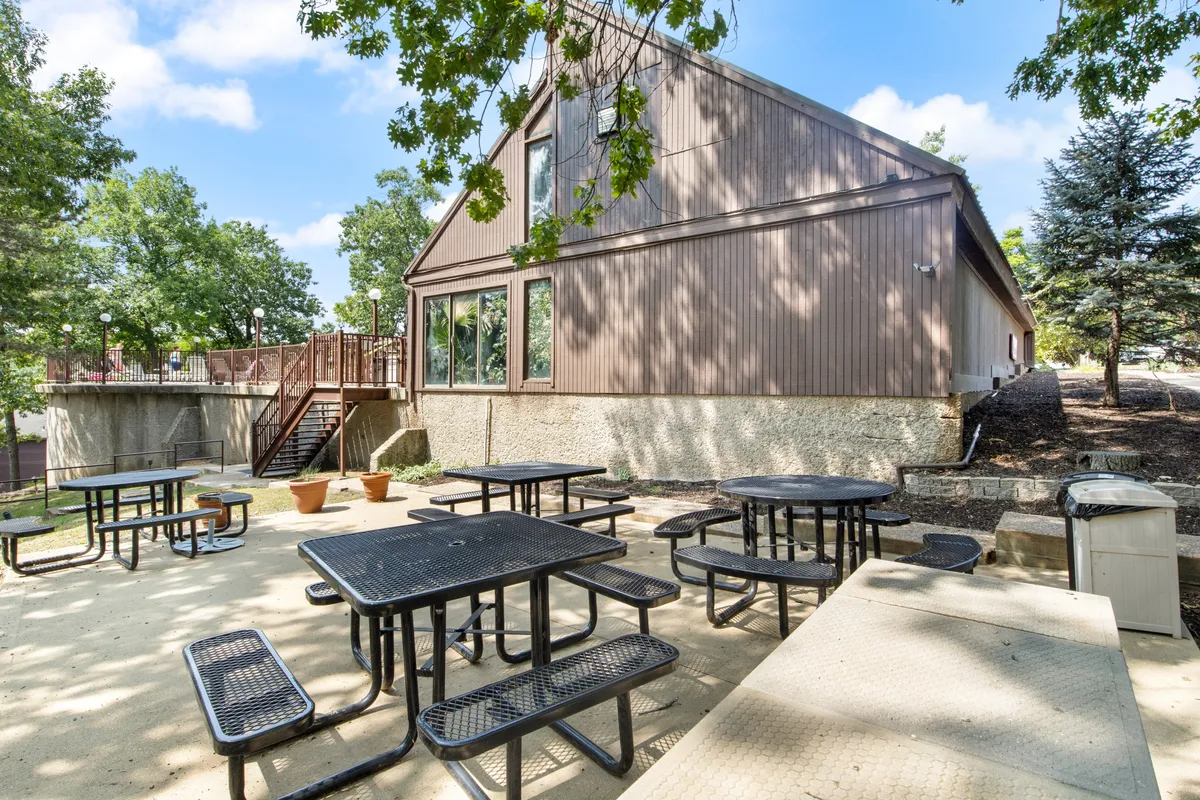 This exterior image captures picnic tables set against the building facade, with tall trees providing natural shade. The inviting setup suggests opportunities for outdoor dining, group gatherings, and casual relaxation under the open sky.